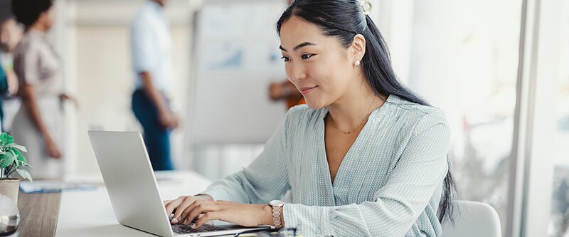 Asian woman working on laptop. Businesswoman busy working on laptop computer at office with colleagues in the background.