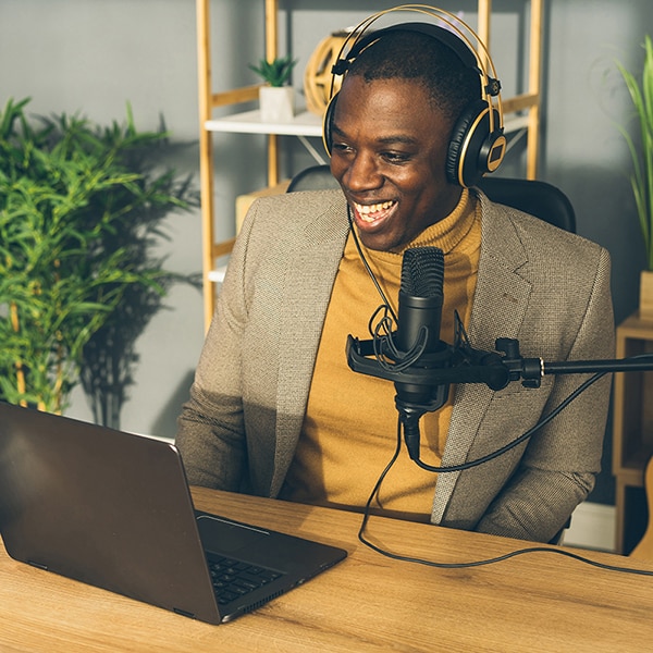 A smartly dressed African-American man sitting at a desk with a laptop, microphone and headset smiling as he works in a bright modern room